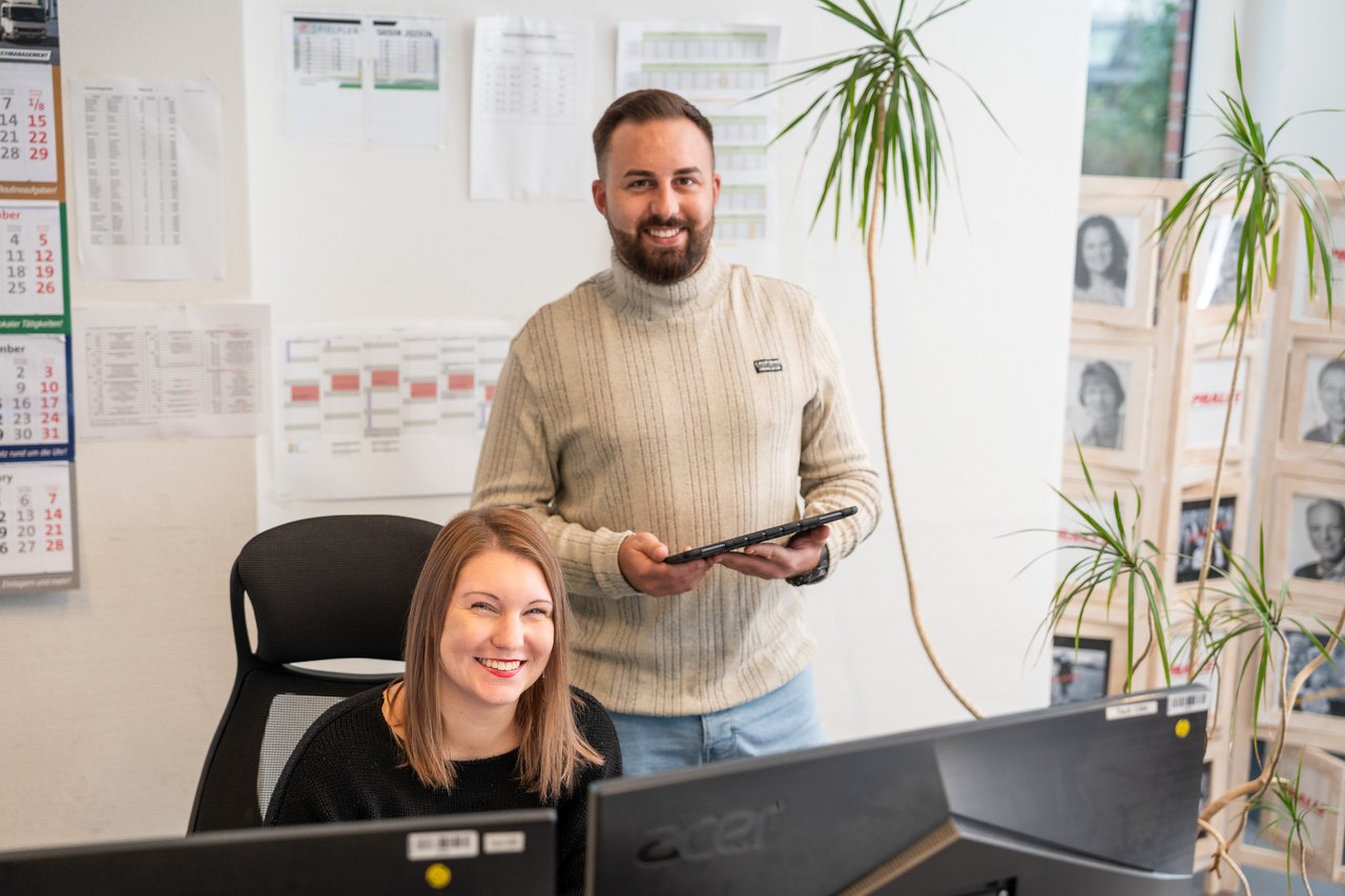 Zwei lächelnde Kollegen im Büro, eine Frau sitzt am Schreibtisch vor Monitoren, ein Mann steht mit Tablet.