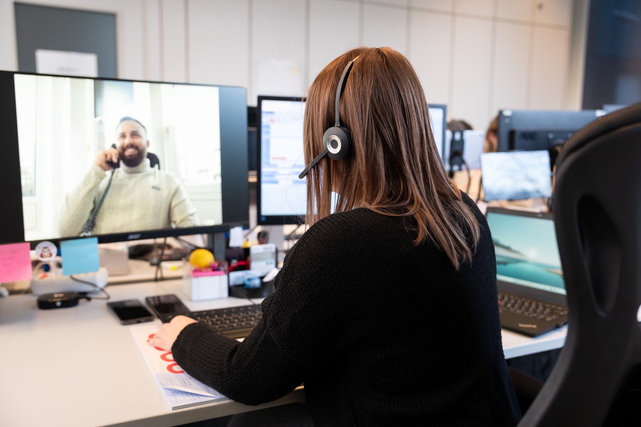 Frau mit Headset am Schreibtisch vor zwei Monitoren; auf dem linken Monitor ist ein lächelnder Mann zu sehen.