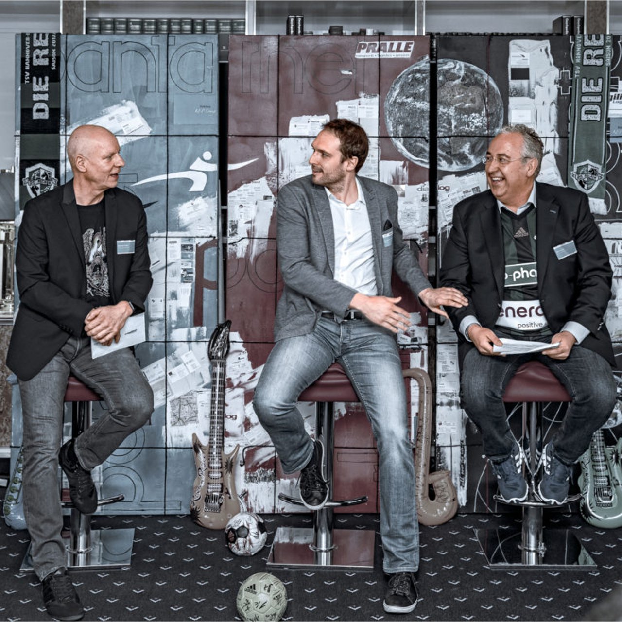 Drei Männer sitzen auf Barhockern vor einer bunten Wand und lachen; E-Gitarren und Fußbälle liegen auf dem Boden.